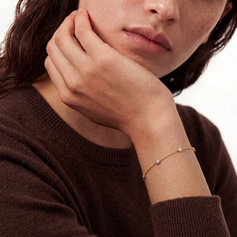Model Image - A close-up lifestyle shot of a model wearing a delicate gold chain bracelet with small clear gemstones on the wrist, paired with a brown knit sweater against a clean white background.