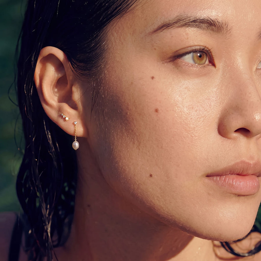 Model Image - A close-up of a woman's ear wearing a delicate gold ear cuff with small clear stones and a dangling white pearl earring, shown outdoors with wet hair.