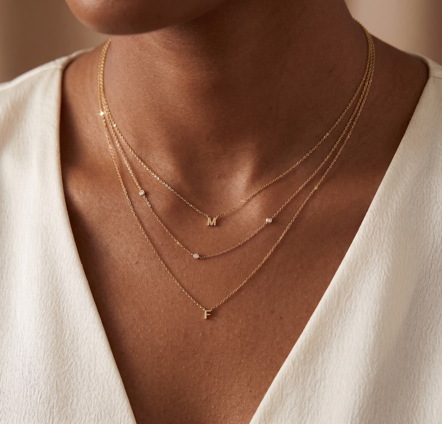 Model Image - A close-up of a model wearing a layered gold necklace set with small letter pendants and tiny clear stones, paired with a white V-neck top.