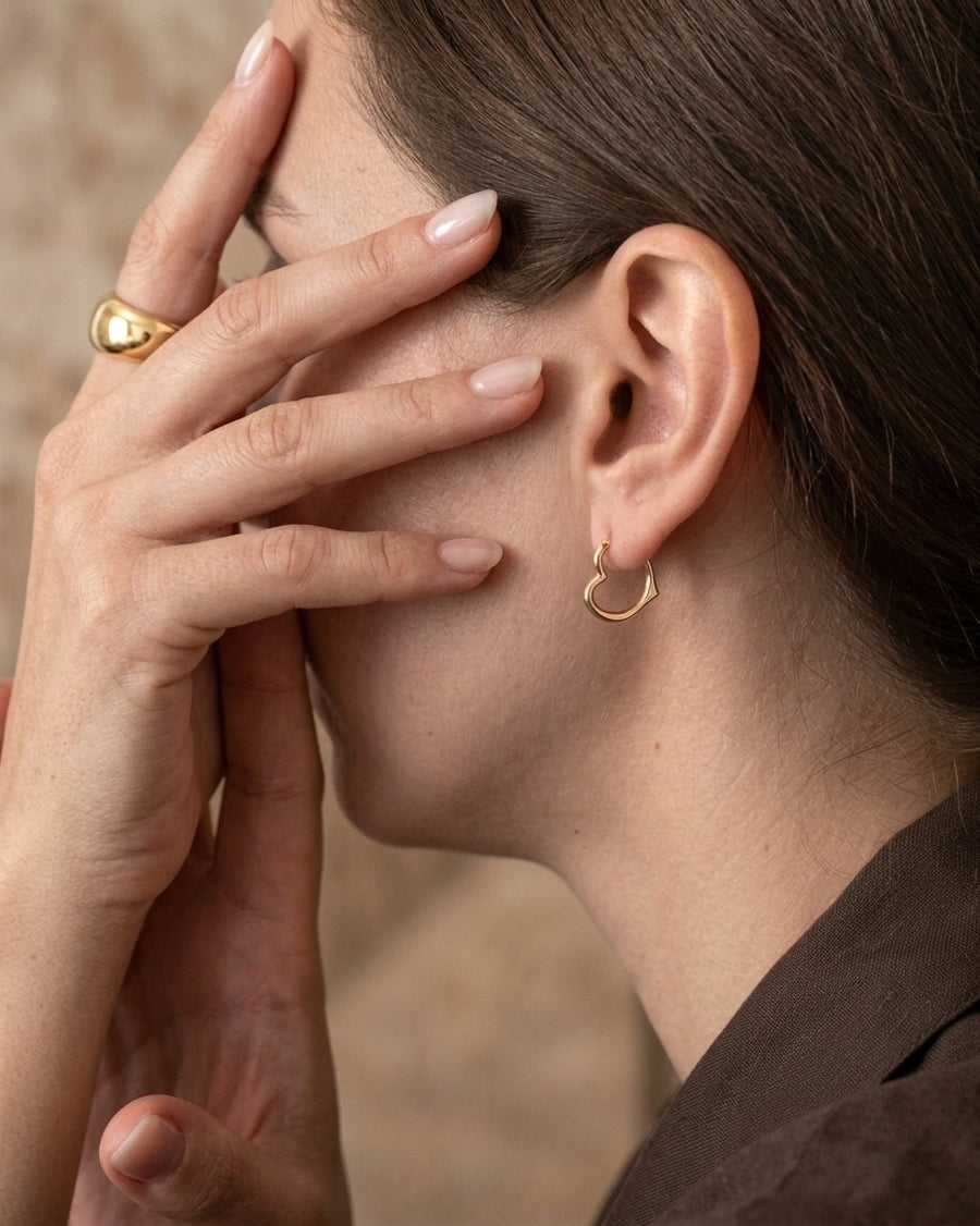 model image - A close-up lifestyle shot of a woman wearing a small gold hoop earring in her ear, with her hand covering part of her face. She is also wearing a gold ring on her finger.