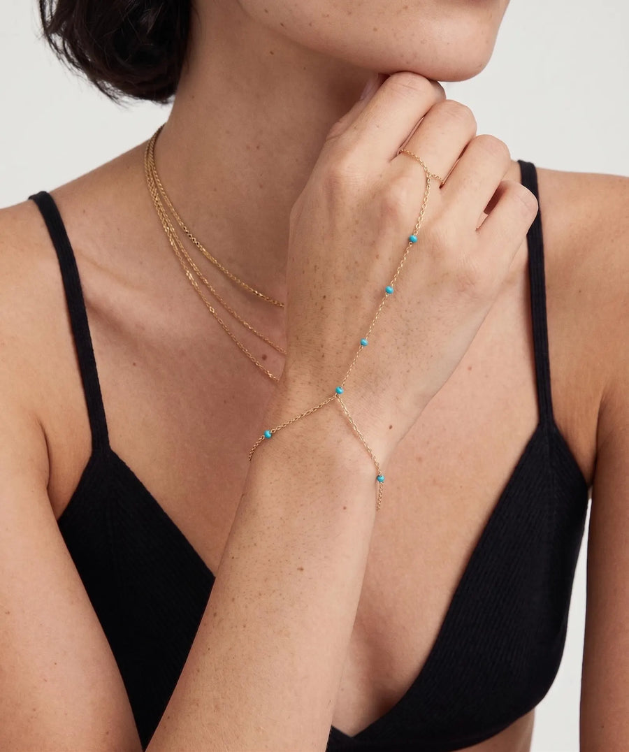 model image - A close-up of a model wearing a delicate gold chain bracelet with small turquoise blue bead accents, styled over a black ribbed camisole top against a light gray background.