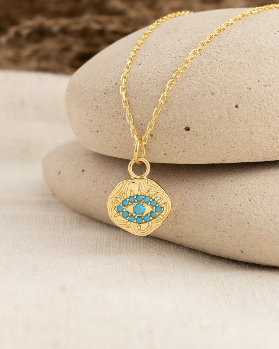 Style Image - 14K gold plated evil eye charm with blue accents shown on a chain against stacked stones.