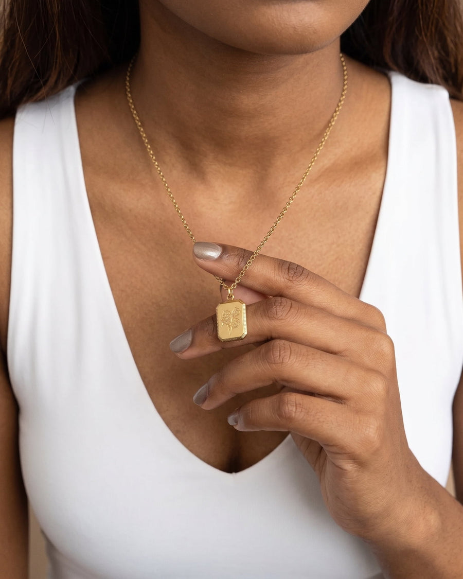 model image - A close-up of a model wearing a delicate gold chain necklace with a small, square, engraved gold pendant, styled with a white sleeveless top.